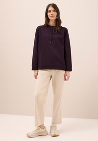 Paarse sweatshirt met een hoge hals, trekkoorddetails en lange mouwen, gecombineerd met beige wijde broek en lichte sneakers.
