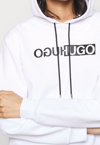 Felpa bianca con un grande logo nero "HUGO", cordini neri e polsini a costine. Tessuto morbido, vestibilità casual, che mette in mostra la parte superiore del torso e le braccia.