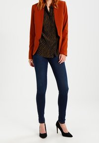 Blazer arancione sopra una blusa a fantasia nera e marrone, abbinato a jeans skinny blu scuro e scarpe con tacco nere.