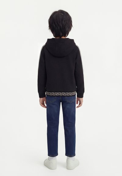 Παιδί που στέκεται με την πλάτη γυρισμένη φορώντας μαύρο φούτερ με κουκούλα και με σχεδιαστή μπορντούρα, μπλε τζιν, λευκές κάλτσες και λευκά αθλητικά παπούτσια σε λευκό φόντο.