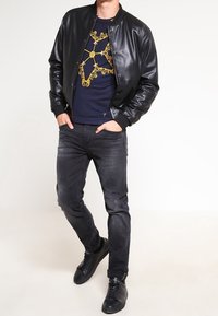 Chaqueta bomber de cuero negro sobre sudadera azul marino con diseño gráfico dorado, combinada con vaqueros oscuros y zapatillas negras.