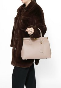 Borsa a mano in pelle beige con forma strutturata, hardware color oro e manico superiore. Indossata con un cappotto di pelliccia marrone.