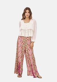 Blusa blanca y texturizada con capas de volantes y botones, combinada con pantalones de pierna ancha con estampado en tonos rosa y marrón que presentan detalles de flecos.