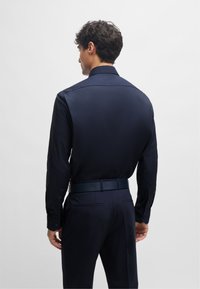 Camicia blu navy in tessuto liscio con colletto strutturato, abbinata a pantaloni scuri e cintura coordinata, vista da dietro.