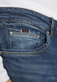 Mörkblå denimjeans med kontrasterande sömmar, med en liten framficka klädd i svarta, vita och röda ränder.