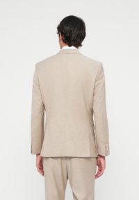 Beige szabott öltönyzakó, strukturált formával, nyakkivágási fülekkel és négy gombos mandzsettával. Simább anyag, finom textúrával és letisztult vonalakkal.