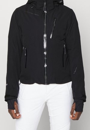 Chaqueta negra con cremallera y múltiples bolsillos con cremallera, agujeros para el pulgar en las mangas, llevada sobre pantalones blancos, mostrada desde el cuello hasta las caderas.