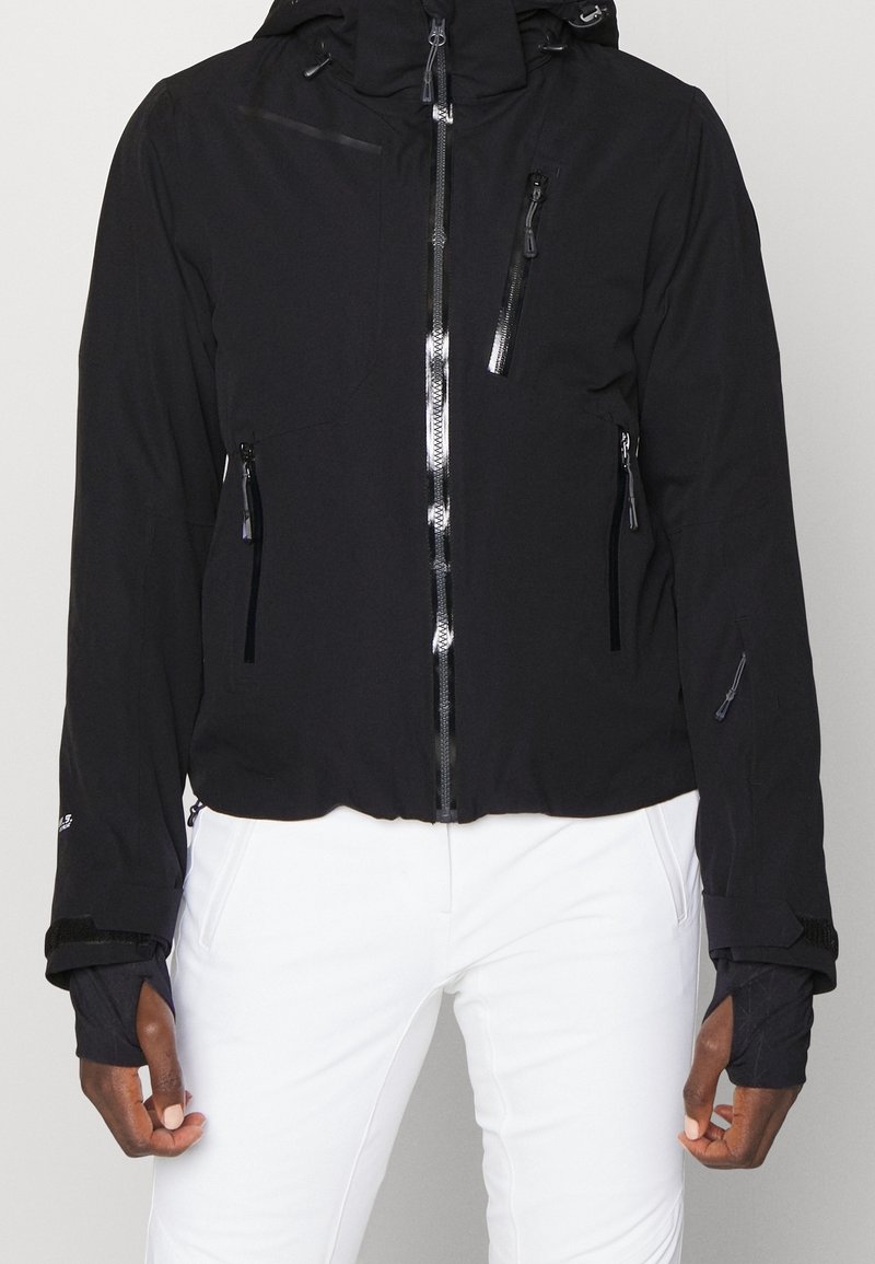 Veste noire zippée avec plusieurs poches zippées, trous pour les pouces dans les manches, portée sur un pantalon blanc, montrée du cou aux hanches.