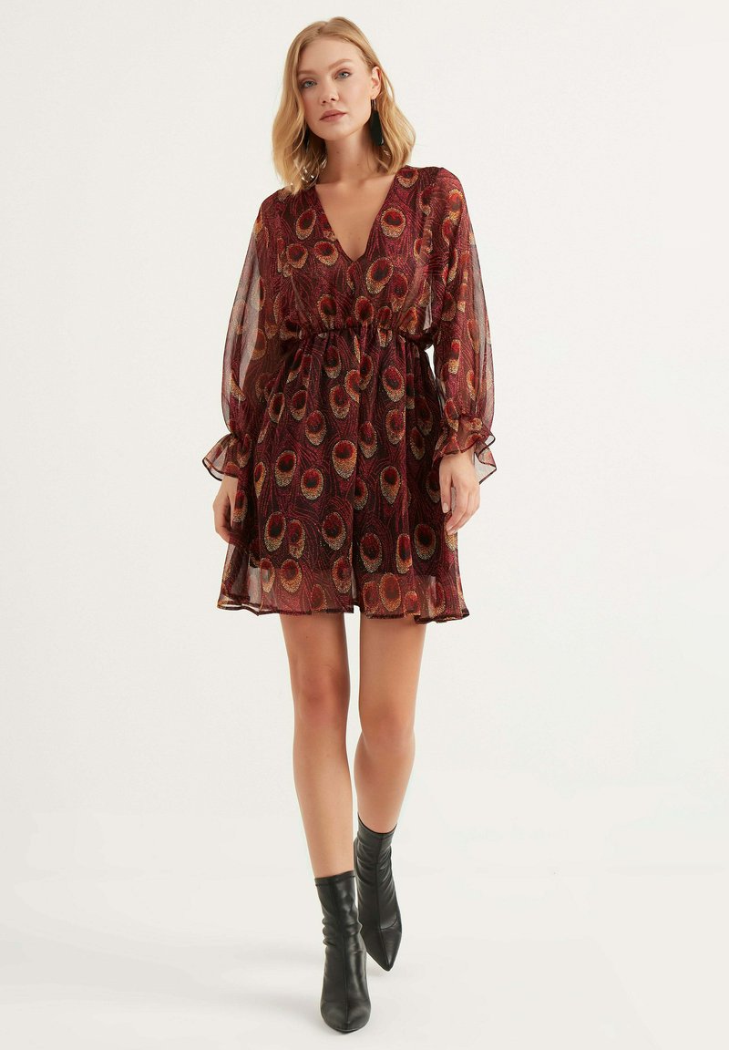 Rotes und golden gemustertes Kleid mit tiefem V-Ausschnitt, langen transparenten Ärmeln, elastischem Bund und weichem, fließendem Stoff. Kombiniert mit schwarzen Stiefeletten.