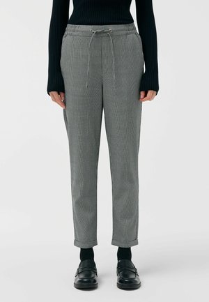 Osoba ubrana w szare spodnie dresowe w kratę ze sznurkiem, czarną prążkowaną bluzkę z długim rękawem, czarne skarpetki i czarne skórzane mokasyny, stojąca prosto.