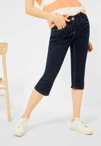 Tummansiniset denim-kesäpökköt, joissa istuva siluetti; edessä nappi, vyölenkit, sivutaskut ja keltaiset sauma-aksentit.