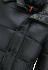 Zwarte pufferjas met een gewatteerd ontwerp, voorzien van een hoge kraag, drukknopen en een oranje trekkoord. Gladde, synthetische textuur.