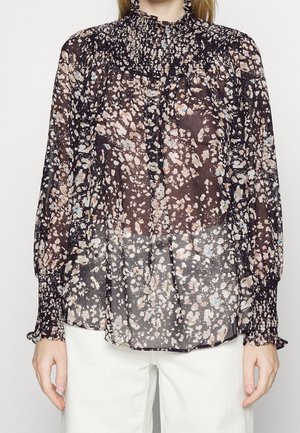 Kvinde iført en gennemsigtig sort bluse med beige og blå abstrakte pletter, smockede manchetter og krave, kombineret med hvide bukser.