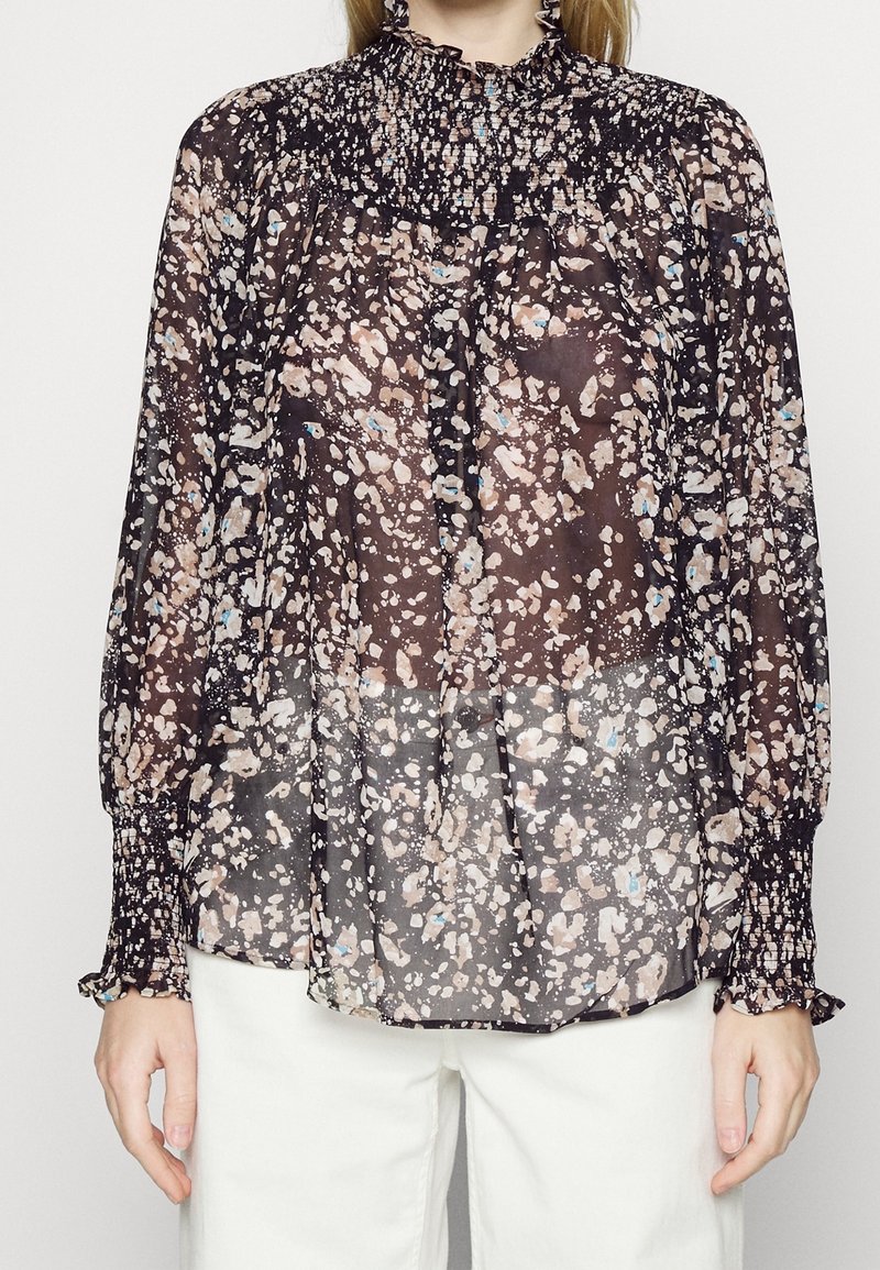 Femme portant un chemisier noir transparent avec des taches abstraites beige et bleue, poignets et col smockés, associé à un pantalon blanc.