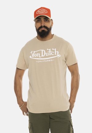 Homme barbu portant une casquette rouge Von Dutch, un t-shirt beige Von Dutch California et un short cargo vert, debout avec les bras détendus le long du corps.