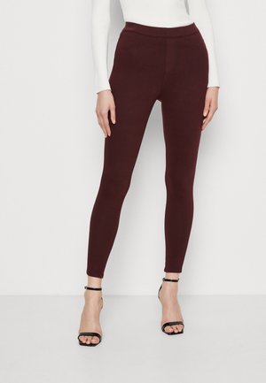 Kvinde iført mørkerøde bukser med høj talje og tætsiddende pasform, kombineret med sorte åbne hæle og en hvid langærmet top, stående mod en hvid baggrund.