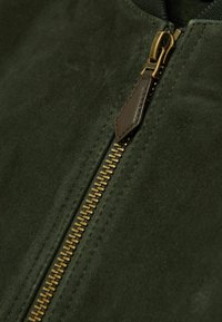 Jachetă din suede verde măsliniu, cu un fermoar din alamă și un tab de piele maro. Textură netedă și un luciu subtil.