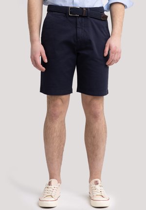 Marineblaue Baumwollshorts mit einer Vordertasche und umgeschlagenen Säumen, kombiniert mit einem geflochtenen braunen Gürtel. Beigefarbene Sneakers mit roten Akzenten runden den Look ab.