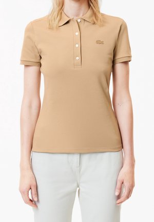 Vrouw die een beige poloshirt met korte mouwen draagt met vier knopen en een klein krokodillenlogo op de linkerborst, gecombineerd met een lichte broek.