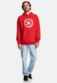 Red hooded sweatshirt with a large white graphic logo in the center, front pocket, ribbed cuffs, and relaxed fit; paired with light blue jeans and black shoes.