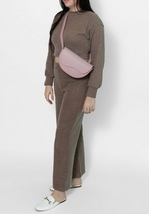 Vrouw die een bruin gebreid loungewear-set draagt met een roze crossbodytas en witte instaploafer, staand tegen een witte achtergrond.