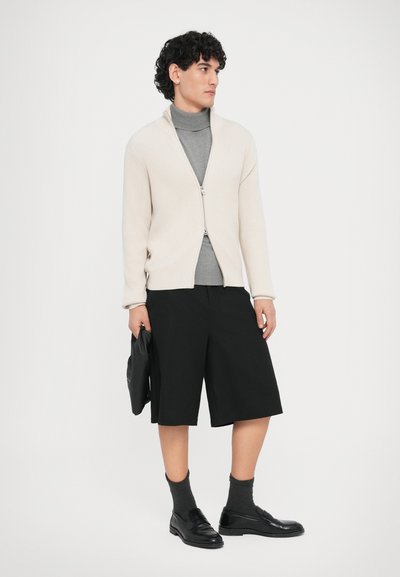 Μπεζ ριγωτό πλεκτό ζακέτα με φερμουάρ, γκρι ζιβάγκο, μαύρο σορτς, γκρι κάλτσες και μαύρα παπούτσια, κρατώντας μια μαύρη τσάντα.