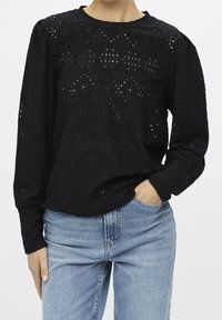 Blouse à manches longues noire à motifs avec découpes complexes et encolure ronde, portée avec un jean bleu clair. Tissu texturé.