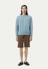 Hellblauer Pullover mit Rundhalsausschnitt und langen Ärmeln; braune Shorts; schwarze Slipper. Das Material sieht weich aus und hat eine strukturierte Oberfläche.