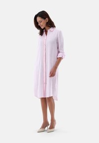 Abito camicia di lino rosa chiaro con chiusura a bottoni, rifiniture in pesca a contrasto, maniche arrotolate e orlo curvo. Indossato con tacchi alti beige.