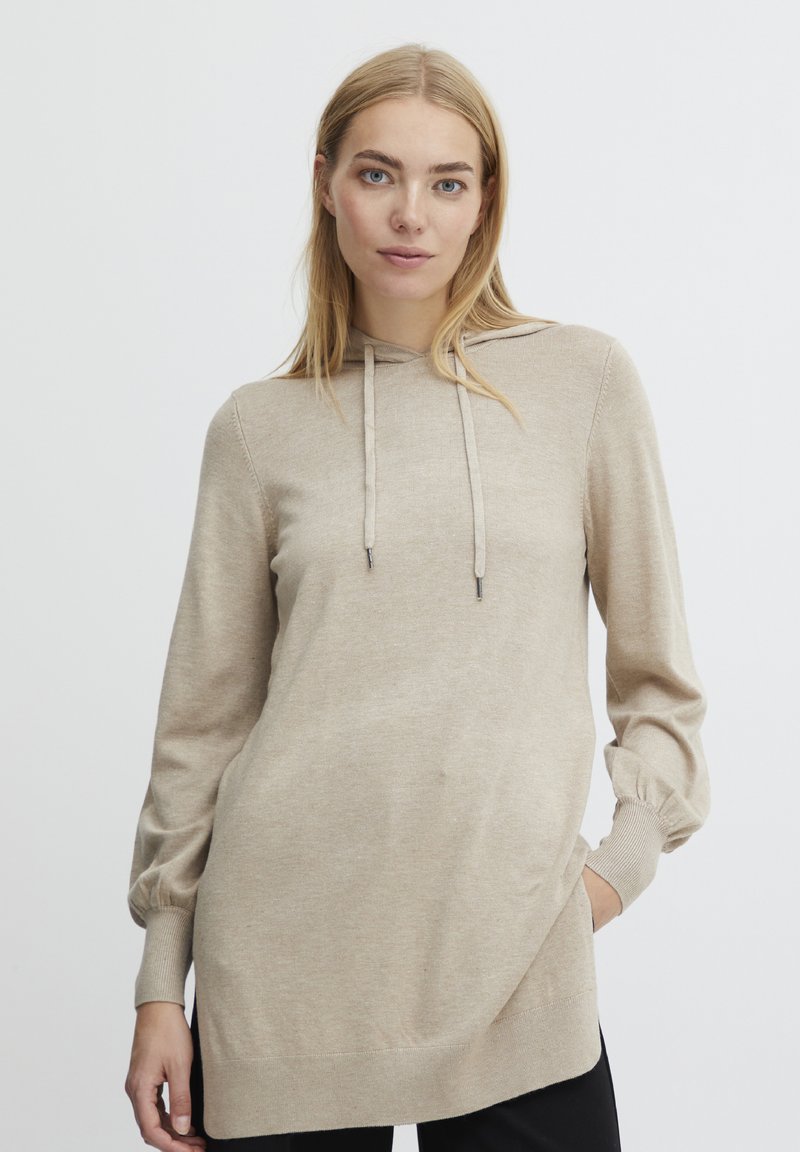 Pull en tricot beige avec capuche, manches longues et poignets ajustés. Texture lisse, coupe décontractée et ourlet arrière légèrement plus long.