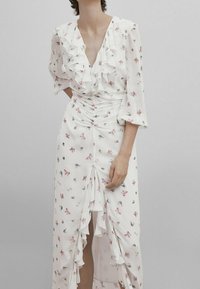 Robe florale blanche avec un col à volants, des manches longues et un devant froncé. Elle présente de petits motifs de fleurs roses et bleues ainsi qu'une fente à l'avant.