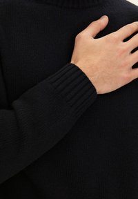 Maglione lavorato a maglia blu navy con collo a imbuto e polsini a costine. La texture è morbida e fine, con un primo piano del maniche e della mano.