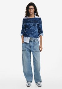 Blauer, strukturierter Off-Shoulder-Pullover, der über einem hellblauen Hemd mit Knöpfen getragen wird, kombiniert mit hoch taillierten, locker sitzenden, hellblauen Jeans und gemusterten Sneakers.