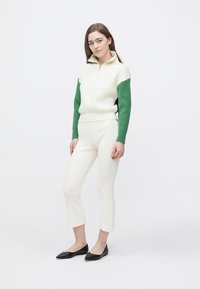 Pull-over blanc à côtes avec manches vertes, col à demi-fermeture éclair. Associé à un pantalon blanc ajusté et des ballerines noires à bouts pointus. Texture lisse.