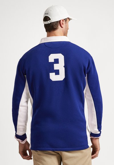 Άνδρας που φορά μπλε και λευκό μακρυμάνικο ράγκμπι πουκάμισο με τον αριθμό 3 στην πλάτη και λευκό καπέλο πόλο, στέκεται με την πλάτη προς τα εμπρός σε απλό φόντο.
