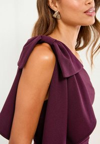 Burgundská off-shoulder šaty s veľkou mašľou na rukáve, s hladkým materiálom a priliehavým strihom. Viditeľné zlaté geometrické náušnice.