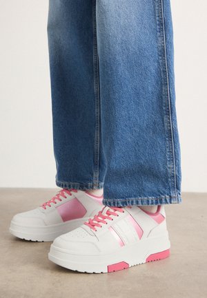 Białe sneakersy z różowymi akcentami, mające gładkie skórzane wykończenie, okrągły czubek i grubą podeszwę, zestawione z niebieskimi dżinsami o szerokich nogawkach.