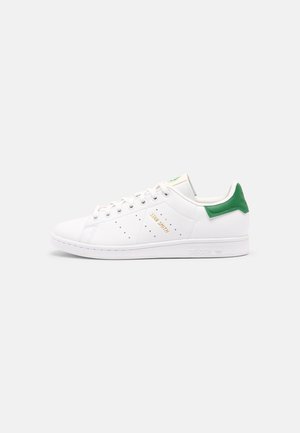 Białe niskie sneakersy Adidas Stan Smith z zielonym językiem z tyłu, perforowanymi paskami po bokach i złotym napisem "Stan Smith" po boku.