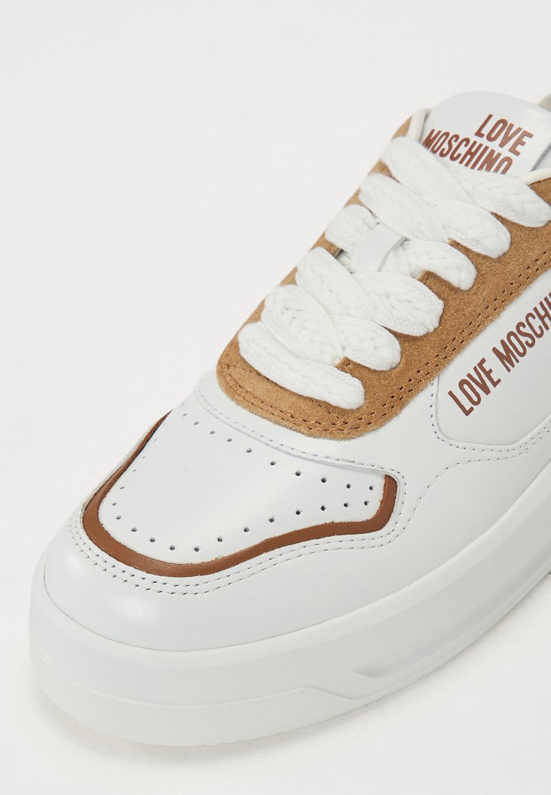 Sneaker in pelle bianca con accenti in suede marrone, caratterizzata da spesse stringhe bianche, perforazioni e il marchio "LOVE MOSCHINO" sul lato.