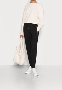 Lichtbeige sweatshirt, zwarte joggingsbroek met strik taille, en witte sneakers. Model houdt een lichtbeige tas vast. Eenvoudig ontwerp, relaxte pasvorm.