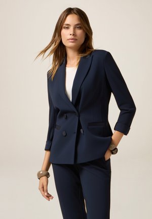 Navy blue tailored double-breasted blazer with a smooth texture, featuring two front pockets and three-quarter sleeves.