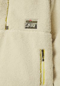 Chaqueta de forro polar color crema con una etiqueta negra y amarilla, un bolsillo con cremallera y detalles amarillos, y una superficie texturizada y suave.