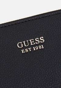 Marineblaues, strukturiertes Lederportemonnaie mit goldfarbenem "GUESS EST 1981" Logo-Detail. Verfügt über sichtbare Ziernähte entlang der Ränder.