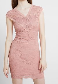 Robe en dentelle rose avec un décolleté en V et des manches courtes, ornée de motifs floraux et d'une coupe ajustée. Texture lisse, silhouette à hauteur de genoux.