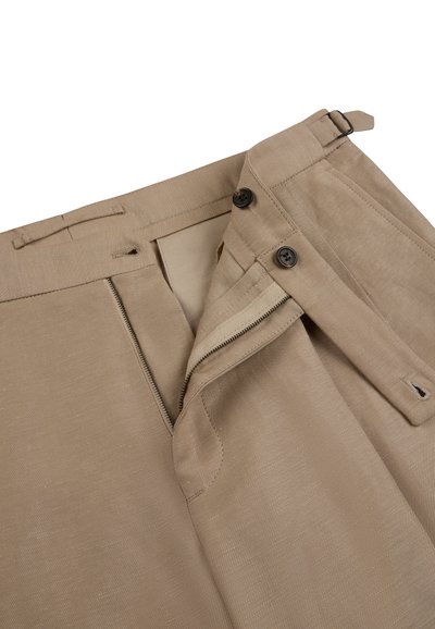 Close-up of beige pants with a partially open zipper, two black buttons, and adjustable side strap on a white background.