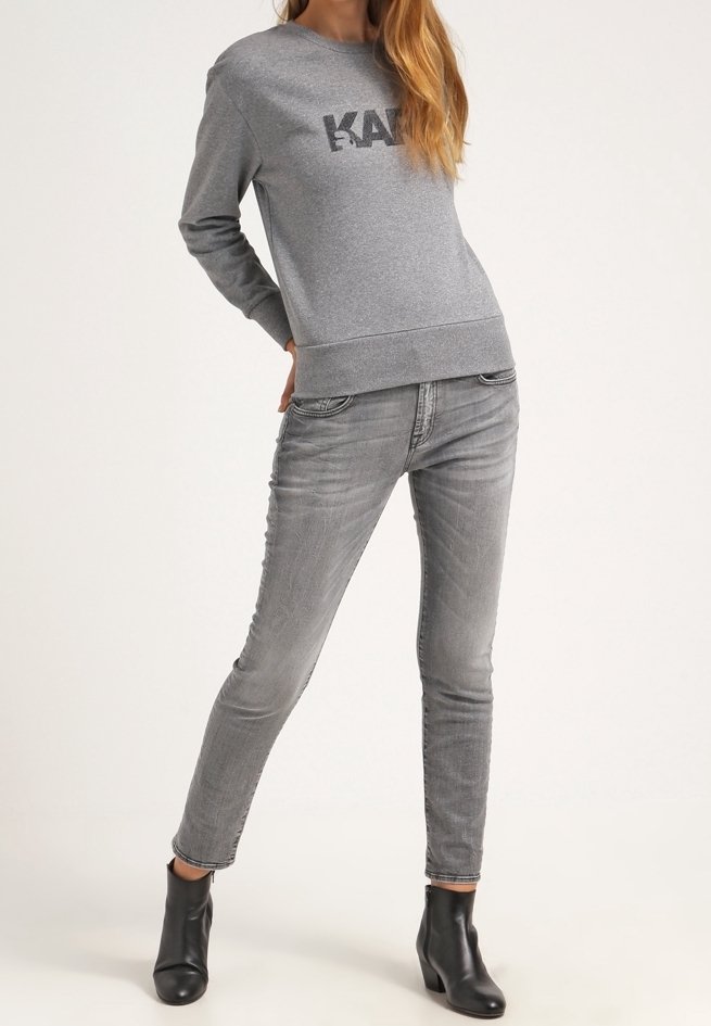 Femme portant un sweat gris avec l'inscription "KARL", jean slim gris et bottines noires, posant les mains dans le dos.