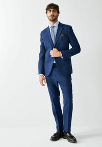 Tengerészkék öltöny vékony szabással, egysoros zakóval és hozzáillő nadrággal. Világoskék ing, mintás nyakkendő és barna díszzsebkendő. Fekete cipő.