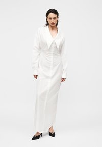 Vrouw draagt een lange witte jurk met knopen, een oversized puntige kraag en zwarte puntige hakken, staand tegen een eenvoudige achtergrond.