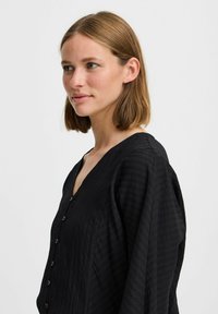 Black button-up shirt with a V-neck, featuring textured fabric and long, flowing sleeves. Subtle vertical stripes visible on the material.