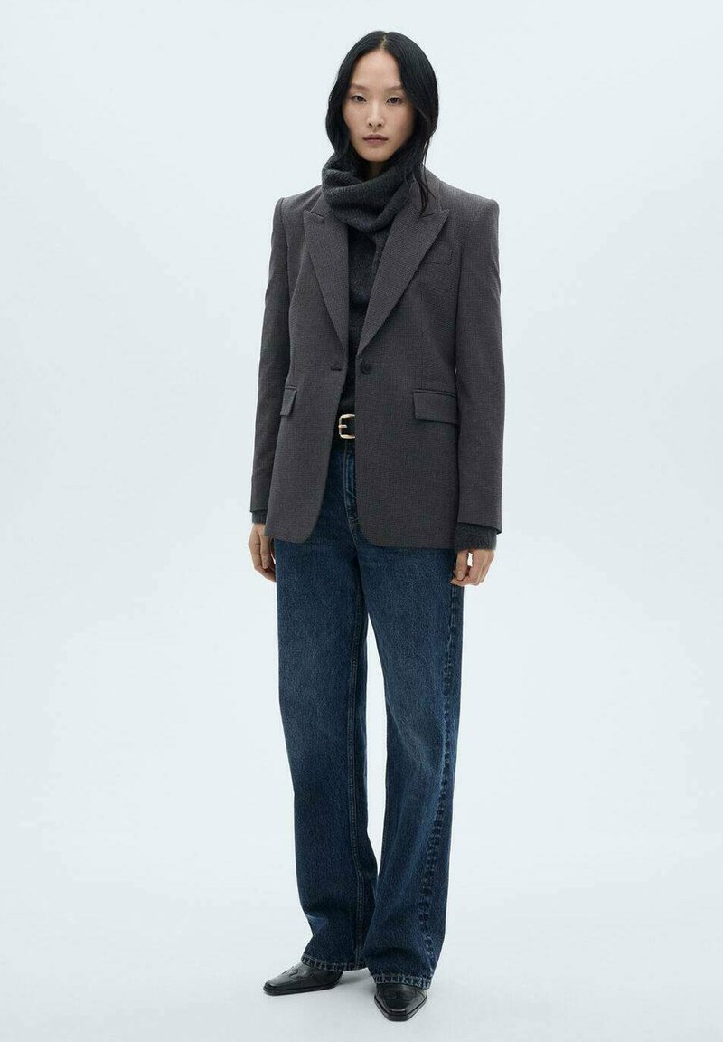 Grå kavaj med breda slag över en mörk polotröja, kombinerat med lösa mörkblå jeans och svarta skor. Slät tyg och strukturerad design.